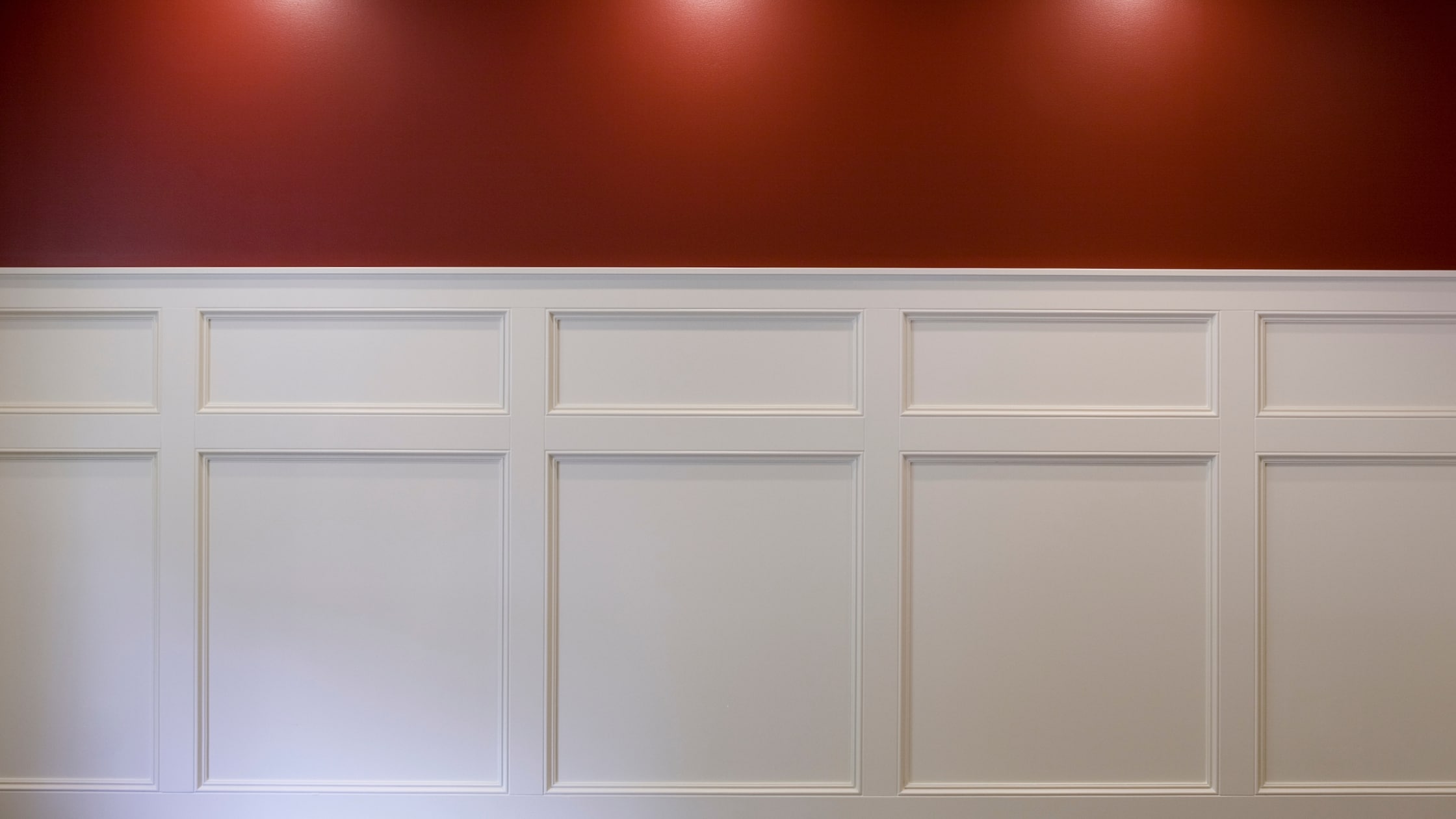 A close-up view of a white wainscotting accent wall with a bold red upper wall and overhead lighting.